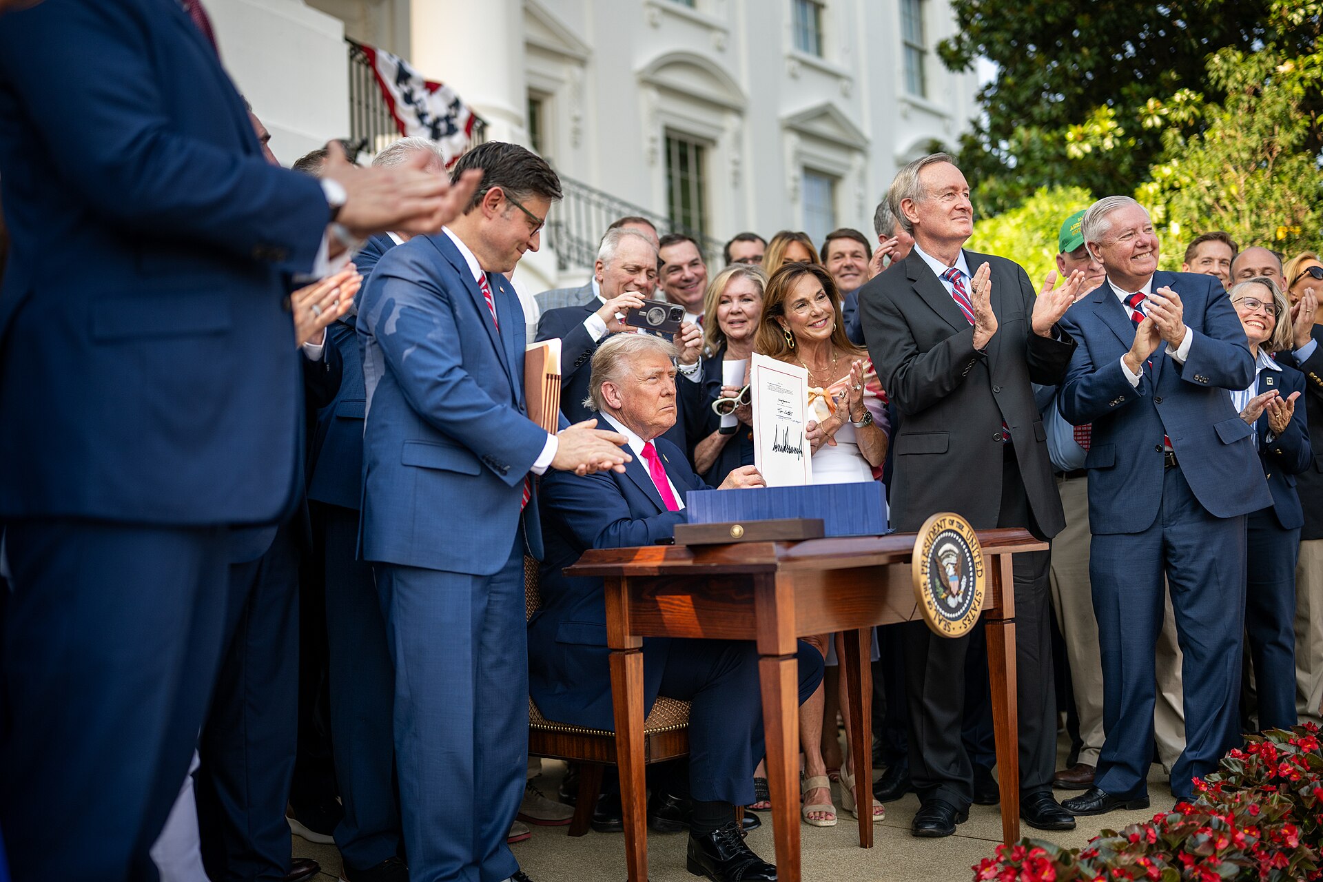 President Trump holds up the signed One Big Beautiful Bill Act, surrounded by applauding legislators on the White House South Lawn